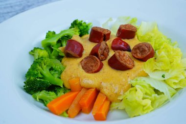 Beyaz tabakta pişmiş brokoli, lahana, havuç ve kremalı sarı sosun üzerinde dilimlenmiş sosisler var..