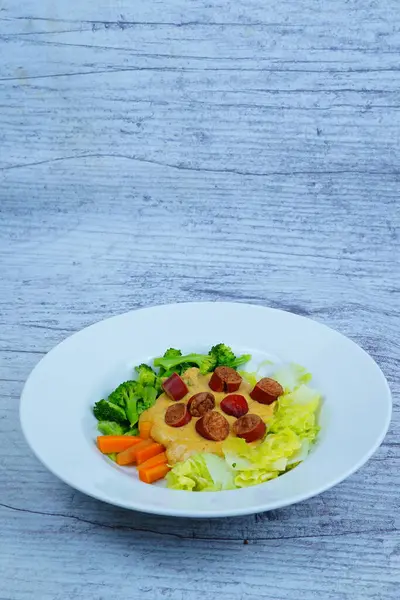 Beyaz tabakta pişmiş brokoli, lahana, havuç ve kremalı sarı sosun üzerinde dilimlenmiş sosisler var..