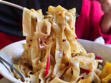 Yakın çekim geniş, düz erişteleri çatalla kaldırır, parlak dokularını ve kırmızı biber parçalarını gösterir, lezzetli bir yemeği işaret eder..