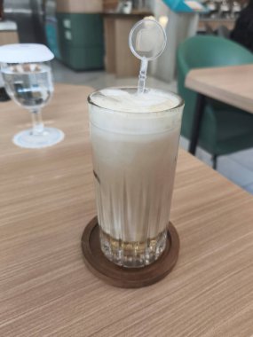 Köpüklü buzlu bir kahve içeceği ve karıştırıcı ahşap bir bardak suyun yanında, ahşap bir masanın üzerinde oturur..