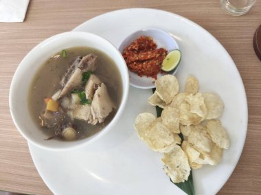 Beyaz tabakta bir kase Sop Buntut ya da Endonezya sığır kuyruğu çorbası var. Yanında da kırmızı biber ezmesi, limon ve ahşap masada kraker var..