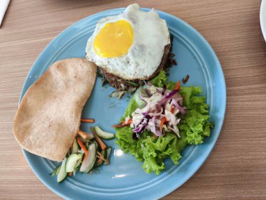 Mavi tabakta Endonezya usulü kızarmış pilav, yumurta, büyük bir karides krakeri, sebze turşusu ve ahşap bir masada taze salata var..