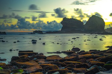 Gün batımında Papuma Sahili yakınlarında şelaleye benzer bir kaya oluşumuyla birlikte kayalık bir sahil şeridinin mistik uzun pozlama çekimi. Doğu Java, Endonezya.