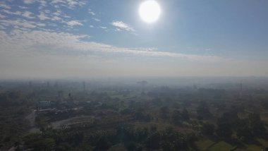 Geniş bir arazinin puslu, güneşli hava manzarası, tarlalar, ağaçlar ve ince bulutlu parlak bir gökyüzünün altında dağınık binalar..