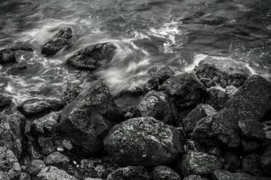Siyah-beyaz, yüksek açılı, uzun açılı bir görüntü dalgalı denizi yakalar. Kıyı boyunca dalgalı kayaları yıkar ve suda bulanık bir hareket yaratır..