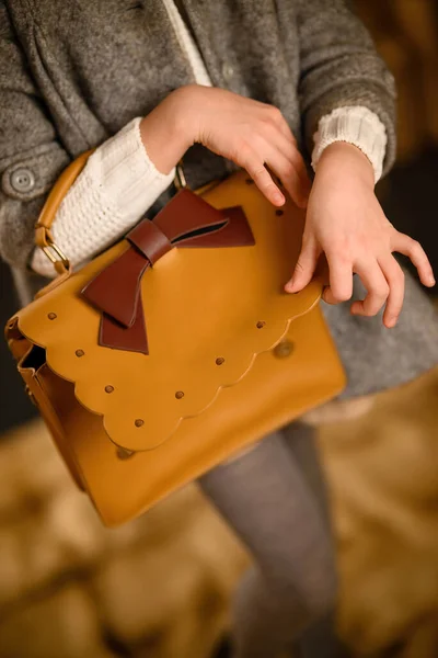 Close-up, girl\'s hand opens yellow leather bag with bow that hangs on second hand of young girl in gray clothes, selective focus on bag, no face. Woman is holding shoulder bag made of genuine soft leather. Women\'s accessories.