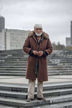Geniş açı, beyaz sakallı, kahverengi kürk süsü ve örgü şapkalı yaşlı bir adamı yakalar. Bulutlu bir gökyüzünün altında kasvetli bir arka planla taş basamaklarda duruyor..