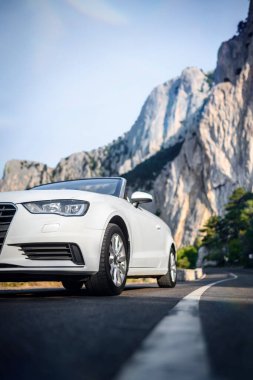 Dönek bir dağ yolunda üstü açık beyaz bir araba, önü kayalık ve arka planda yeşillik. Gün ışığında alçak açılı çekim, dikey oryantasyon, insan yok. Seyahat ve lüks temalar için ideal.