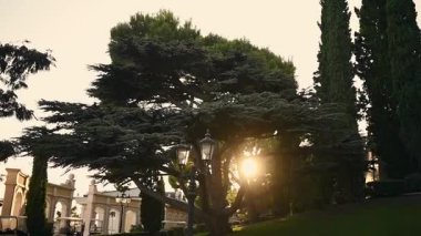 Panoramik çekim sağa doğru hareket ediyor, yemyeşil bahçelerde büyük bir kale arazisi ortaya çıkarıyor. Uzun selvi ağaçları, güneş ışığının filtrelediği sahneyi çerçeveler ve mimari detayları vurgular..