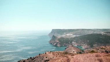 Uçsuz bucaksız okyanusla buluşan engebeli bir kıyı şeridinin panoramik görüntüsü güneşli bir günde yüksek bir noktadan çekilmiş. Kamera sola dönüyor, dramatik uçurumları ve sakin denizi gösteriyor..
