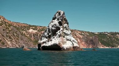 Kaya oluşumu, engebeli kayalıkların ve seyrek bitki örtüsünün kıyı manzarasıyla çevrili turkuaz deniz sularından dramatik bir şekilde yükselir. Açık gökyüzü ile gün ışığı.