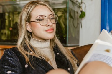 Kitap Günü, 23 Nisan. Gözlüklü sarışın kadın oturma odasında kitap okuyor. Huzur.