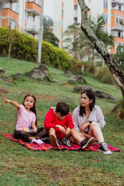 Uluslararası Çocuk Günü. Latin anne dışarıda çocuklarıyla eğleniyor. Yere oturmuş manzarayı işaret ederek oturuyorlar. Aile kavramı. İki küçük çocuğu olan bekar bir anne..