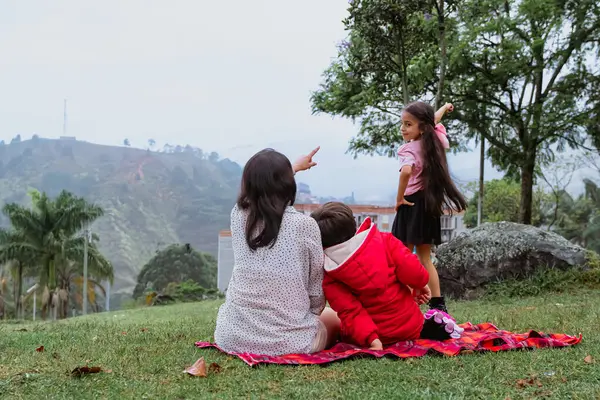 International Children Day. Latina mother having fun with her young children outdoors. Sitting on the ground pointing at the landscape. Concept of family. Single mother with two small children.