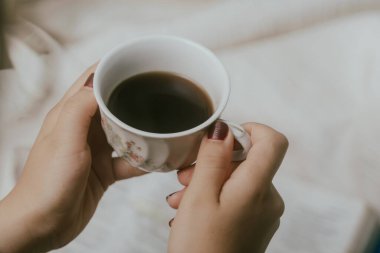 Elinde kahve fincanı ve okumak için yatağında kitap tutan bir kadın. Evde kitap okuyan genç bir kadın. Kapat..