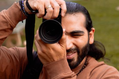 Kahverengi ceketli, uzun saçlı, sakallı Latin bir adam tatilini belgeliyor ve ülkesinin altın saatinin tadını çıkarıyor. Dünya fotoğrafçılık ve seyahat günü.