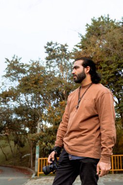 Kahverengi ceketli, uzun saçlı, sakallı Latin bir adam tatilini belgeliyor ve ülkesinin altın saatinin tadını çıkarıyor. Dünya fotoğrafçılık ve seyahat günü.