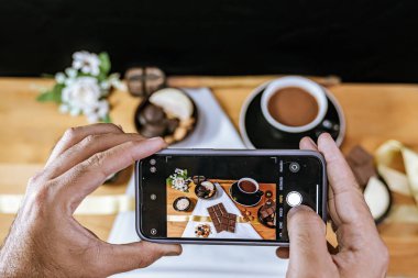 Elinde cep telefonu olan bir adam çikolata işinin fotoğrafını çekiyor. Uluslararası çikolata günü, iş ve finans. Siyah beyaz çikolata..