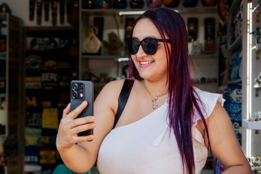 Siyah gözlüklü Latin bir kadın cep telefonuyla konuşurken seyahat edip aksesuar alıyor. Anılar. İnsanlar gülümsüyor.