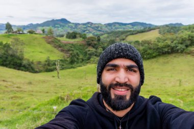 Siyah sakallı yetişkin Latin bir adam Medellin, Kolombiya yakınlarındaki dağlarda selfie çekiyor ve gülümsüyor..