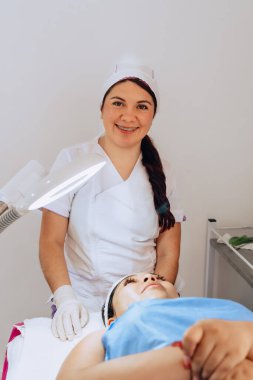 Güzellik merkezindeki iki genç Kolombiyalı kadının fotoğrafı. Dermatolog kameraya bakıyor ve müşterisine yüz temizleme ameliyatında ne yapacağını anlatıyor..