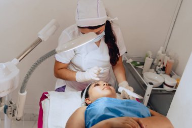 Güzellik merkezindeki bir güzellik uzmanının, yüz temizleme tedavisini tamamlamak için genç melez bir Kolombiyalı kadının yüzüne güneş kremi sürerken fotoğrafı..