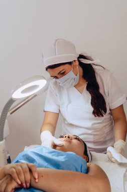 Güzellik merkezindeki bir güzellik uzmanının, yüz temizleme tedavisini tamamlamak için genç melez bir Kolombiyalı kadının yüzüne güneş kremi sürerken fotoğrafı..