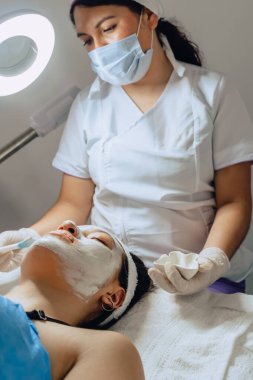 Yüz temizleme kliniğinde genç bir Kolombiyalı Latin kadının yüzüne çamur maskesi takan kadın dermatolog fotoğrafı..