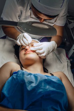 Dermatolog ellerinin sivilceleri ve sivilceleri temizlerken çekilmiş fotoğrafı. Klinikte lambalı ve büyüteçli Kolombiyalı Latin bir kızın sivilce temizleme aleti. Yüz bakımı, cilt bakımı..