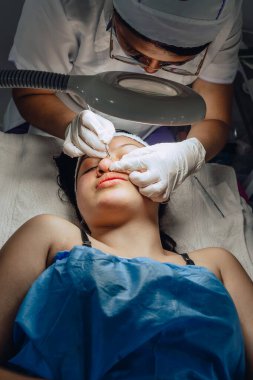 Dermatolog ellerinin sivilceleri ve sivilceleri temizlerken çekilmiş fotoğrafı. Klinikte lambalı ve büyüteçli Kolombiyalı Latin bir kızın sivilce temizleme aleti. Yüz bakımı, cilt bakımı..