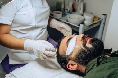 Sakallı, mestizalı, nemlendirici jeli ve kapalı gözleri olan yetişkin bir erkek için profesyonel deri yüzme, nemlendirici ve yüz temizleme fotoğrafı..