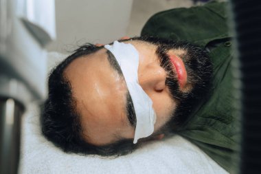 Sakallı, mestizalı, nemlendirici jeli ve kapalı gözleri olan yetişkin bir erkek için profesyonel deri yüzme, nemlendirici ve yüz temizleme fotoğrafı..