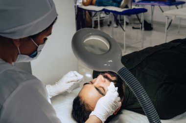 Cildiyecinin, sakallı Kolombiyalı mestizo yetişkin bir erkeğin yüzündeki nemlendirici jeli temizlerken çekilmiş resmi. Profesyonel bir cilt soyma, nemlendirici ve yüz temizleme tedavisi sırasında..