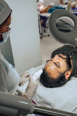 Sakallı, melez bir Kolombiyalı adamın güzellik merkezindeki fotoğrafı. Yüz temizleme operasyonu yapılıyor ve dermatolog yüz mikro akımı cihazıyla arınma aşamasına geçiyor..