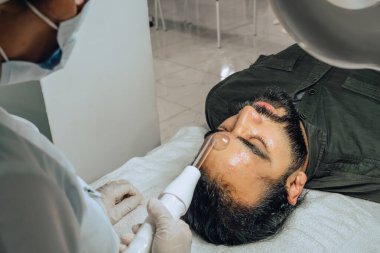Sakallı, melez bir Kolombiyalı adamın güzellik merkezindeki fotoğrafı. Yüz temizleme operasyonu yapılıyor ve dermatolog yüz mikro akımı cihazıyla arınma aşamasına geçiyor..