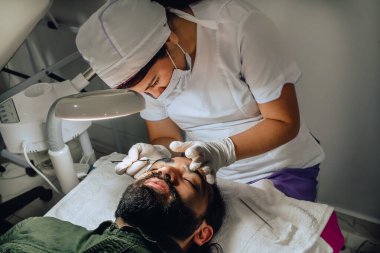 Kadın dermatologların sivilceleri ve sivilceleri temizlerken çekilmiş bir fotoğraf. Sakallı, sakallı, sakallı ve yüz tedavisinde büyüteç kullanan Kolombiyalı bir adamın sivilce temizleme aleti..