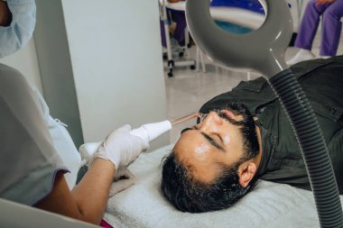 Sakallı, melez bir Kolombiyalı adamın güzellik merkezindeki fotoğrafı. Yüz temizleme operasyonu yapılıyor ve dermatolog yüz mikro akımı cihazıyla arınma aşamasına geçiyor..