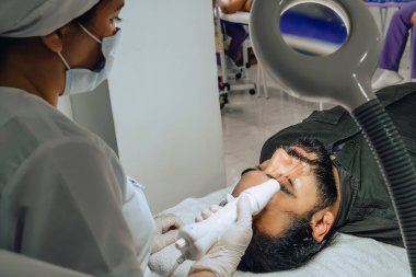 Sakallı, melez bir Kolombiyalı adamın güzellik merkezindeki fotoğrafı. Yüz temizleme operasyonu yapılıyor ve dermatolog yüz mikro akımı cihazıyla arınma aşamasına geçiyor..