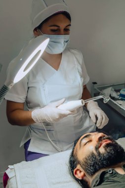 Sakallı, mestizo Kolombiyalı bir adamın güzellik merkezindeki fotoğrafı. Yüz temizleme operasyonu yapılıyor ve dermatolog yüz mikro akımı cihazıyla arınma aşamasına geçiyor..