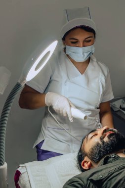 Sakallı, mestizo Kolombiyalı bir adamın güzellik merkezindeki fotoğrafı. Yüz temizleme operasyonu yapılıyor ve dermatolog yüz mikro akımı cihazıyla arınma aşamasına geçiyor..