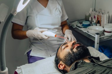 Sakallı, mestizo Kolombiyalı bir adamın güzellik merkezindeki fotoğrafı. Yüz temizleme operasyonu yapılıyor ve dermatolog yüz mikro akımı cihazıyla arınma aşamasına geçiyor..
