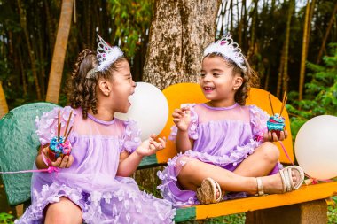 Kolombiyalı ikiz kardeşler lavanta rengi elbiseler içinde yeşil bir parkta bankta otururken ellerinde kekler ve beyaz balonlar tutuyorlar.