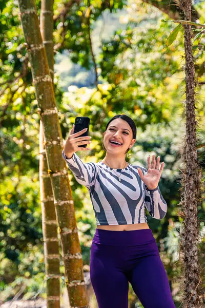 Santuario, Risaralda, Kolombiya 'da kırsal oteldeki tropikal bitki örtüsüyle çevrili çizgili uzun kollu gömlekli ve mor taytlı genç bir kadın telefon tutuyor ve elini kaldırıyor.