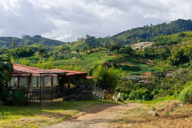 Kolombiya 'nın kırsal kesimindeki kırmızı çatılı bina yeşil teraslar, ağaçlar ve bulutlu gökyüzü ile çevrili.