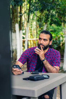 Otuzlu yaşların başında, açık havada oturmuş, akıllı telefonu tutarken mesafeye bakıyor ve bir karar vermeyi düşünüyor.