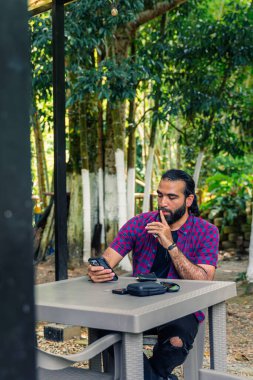 30 'lu yaşların başında bir adam açık havada kırsal masada oturuyor, eli ağıza yakın ve düşünceli bir ifade, elinde akıllı telefonla karar vermeyi düşünüyor.