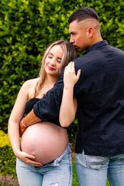 Hamile bir kadın, dış mekan ortamında partnerinin kucakladığı, sakin ve koruyucu bir tavırla ileriye bakarken karnını tutan, sessiz bir bağ ve beklenti anını paylaşan..