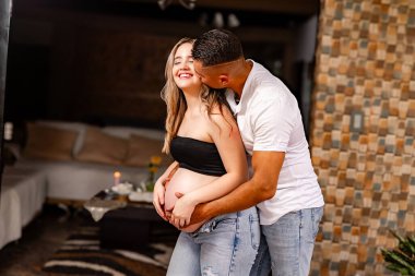 Erkek, hamile kadını kucaklıyor ve öpüyor. Kapalı bir ortamda, o huzurlu bir aşk, şefkat ve duygusal bağ anında gözleri kapalı gülümserken..