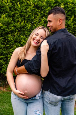 Hamile kadın dışarıda partneriyle duruyor, karnını tutarken koruyucu bir jest ile sarılıyor, duygusal destek ve bağ kurma anını paylaşıyor..