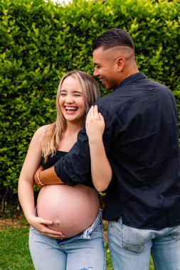 Açık hava ortamında partnerinin kucakladığı hamile bir kadın. Sıcak bir gülümseme ve duygusal bir bağ paylaşıyor. Huzurlu bir aşk ve korunma anında karnını tutuyor..